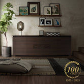 Credenza moderna a quattro porte in finitura moka della collezione Modigliani di Piombini, con intaglio decorativo centrale, ambientata in un soggiorno elegante con quadri di volatili, tappeto tessuto e pianta in vaso. Logo centenario Piombini 1925-2025 in basso a destra.